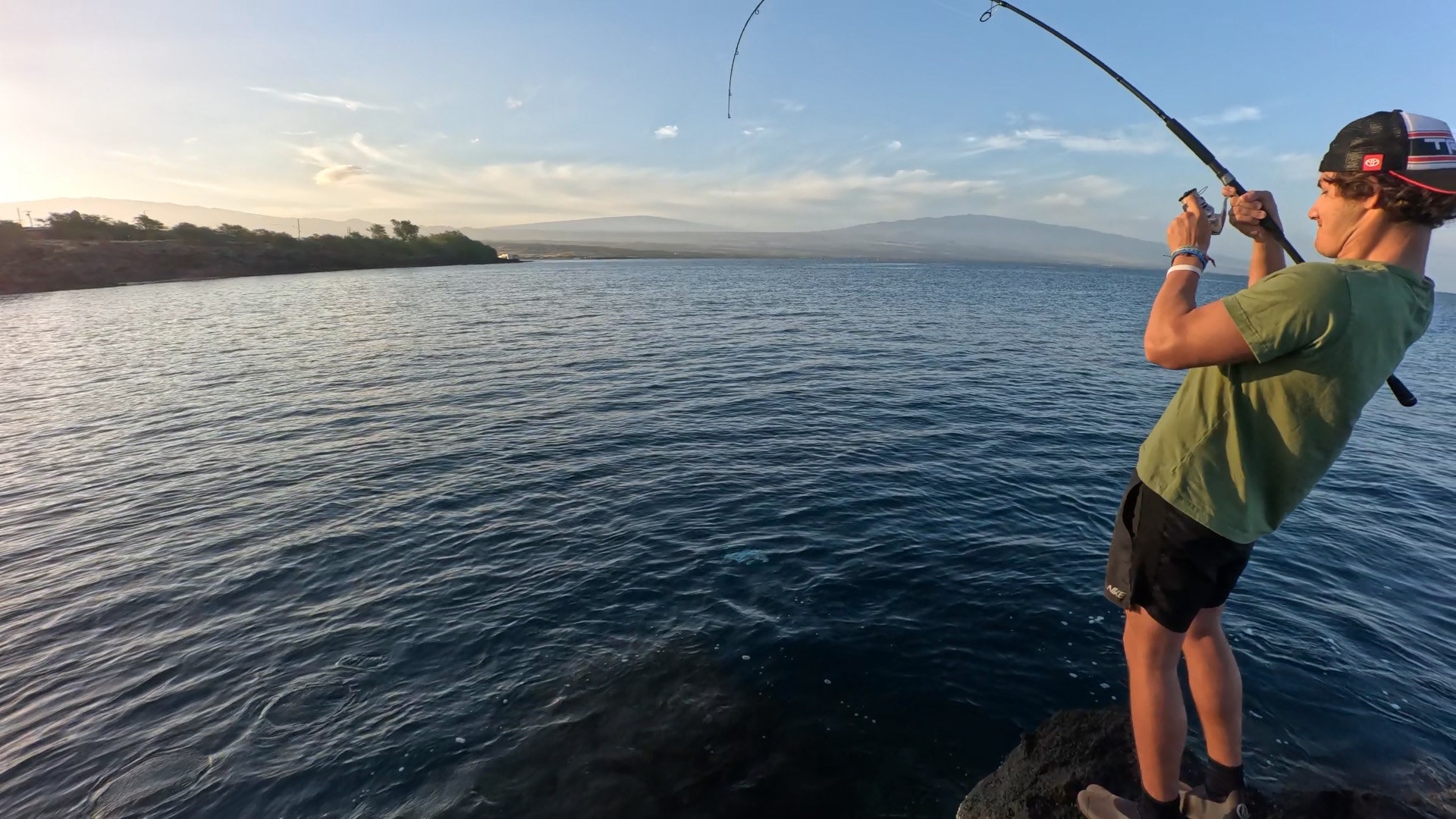 Angler fighting a fish with a bent fishing rod from the rocky shoreline on Hawaii's Big Island.